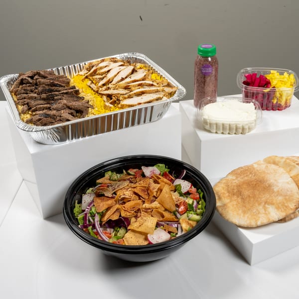 Catering spread with a large tray of rice, beef, and chicken, fattoush salad, pita bread, and sauces.